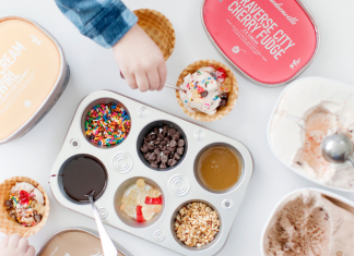 A child's hand celebrating Valentin's Day reaching over a muffin tin full of ice cream toppings surrounded by Hudsonville Ice Cream tubs with an ice cream scooper.
