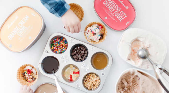 A child's hand celebrating Valentin's Day reaching over a muffin tin full of ice cream toppings surrounded by Hudsonville Ice Cream tubs with an ice cream scooper.